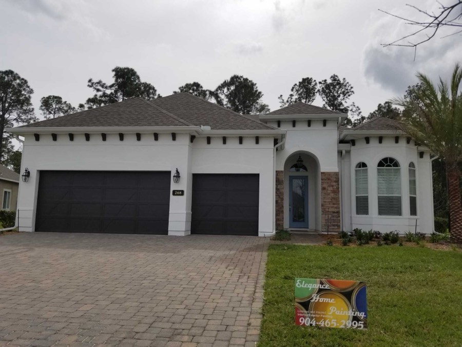 A white stucco home with dark garage doors, freshly painted, with an Elegance Home Painting sign in the front yard