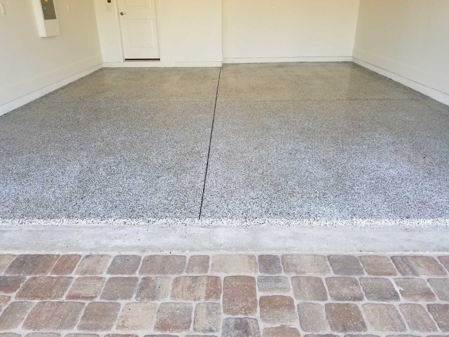 A garage floor finished with grey flake epoxy coating meeting a brick paver apron